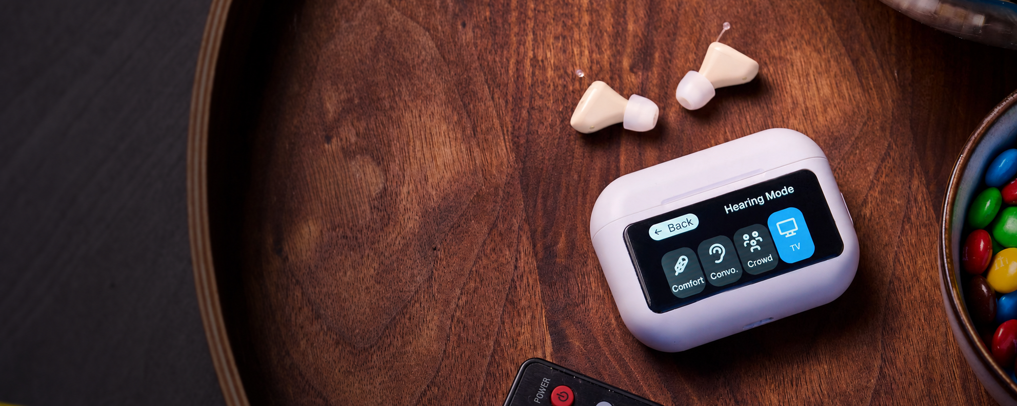 A white and black hearing device with a small display screen, resting on a dark wooden tray next to its charging case, two beige earbuds, and a remote control.